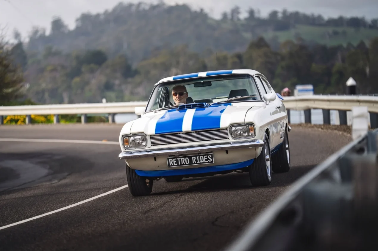White Ford Capri Mk1 with blue racing stripes and driver driving on a curved mountain road with guardrails and "Retro Rides" number plate