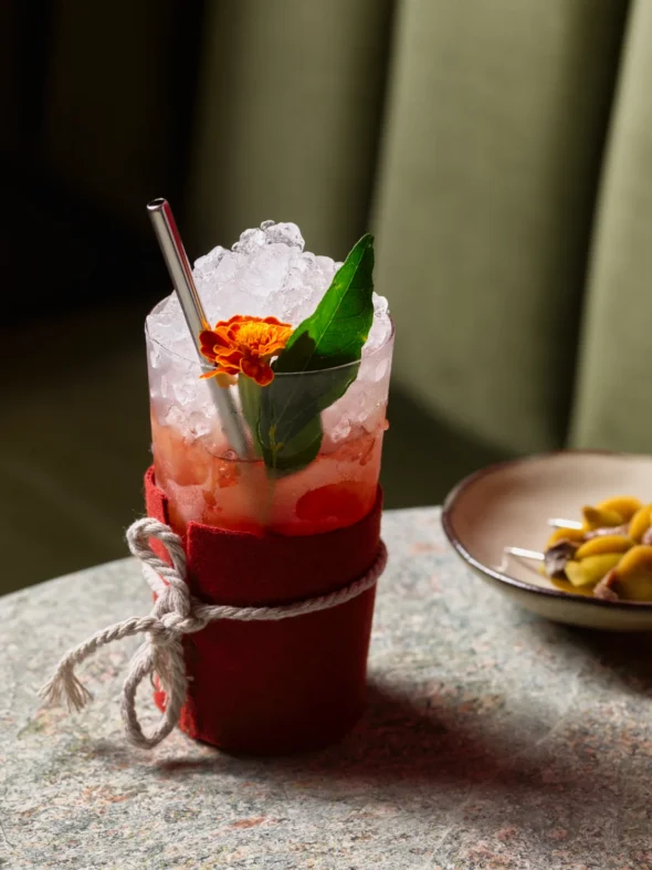 Pink Cashmere cocktail in a red felt-wrapped glass topped with crushed ice, mint leaf, and marigold flower, served with a side of marinated olive skewers at Tailor Room bar